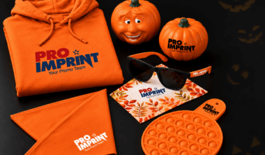 Orange Halloween-themed promotional products including a branded hoodie, pumpkin-shaped stress balls, custom sunglasses with microfiber cloth, a bandana, and a pop-it fidget toy, displayed on a black background with jack-o'-lantern accents.